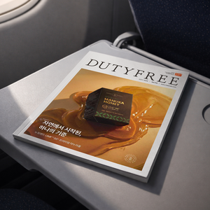 A Duty Free magazine or booklet resting on an airplane tray table, showing a honey-themed cover with a dark box labeled 'Manuka Honey' and Korean text.