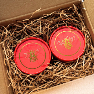 Two round red gift boxes with gold bee designs nestled in brown paper shred inside a cardboard box.