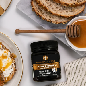 A jar of Manuka Doctor Manuka Honey Monofloral UMF 22+ MGO 970+ placed on a white surface with sliced bread, a honey dipper, and a bowl of honey in a styled flat-lay arrangement.