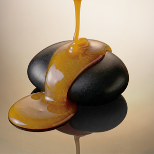 A glossy honey being poured over smooth dark stones on a reflective surface against a warm beige background.