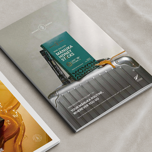 A printed marketing brochure or magazine page for Manuka Honey Sticks lies on a light fabric surface. The page features a silver suitcase graphic with a teal honey stick package shown inside, plus white text reading 'MANUKA HONEY STICKS' and 'YOUR MANUKA ROUTINE, WHEREVER YOU GO.