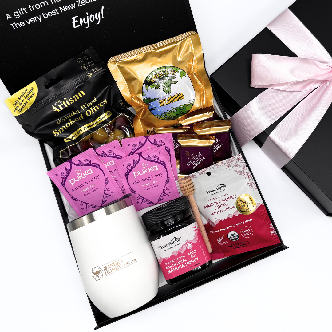 Gift set with various products including a white tumbler, pink packets, and honeycomb, with a black box and pink ribbon on a white background.