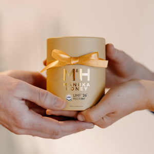 A person's hands holding a gold cylindrical jar of M&H Manuka Honey with a matching gold ribbon bow, set against a soft beige background.