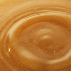 A close-up texture shot of a smooth, glossy golden-brown liquid, likely a cosmetic cream, honey, or oil, with concentric ripples.