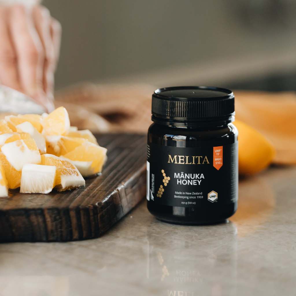 A jar of Melita Mánuka Honey sits on a marble countertop next to a wooden cutting board with sliced oranges and a blurred hand in the background.