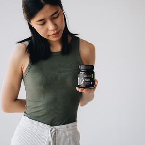 A woman wearing an olive green ribbed tank top and white drawstring pants holds a jar of Melita Manuka Honey against a plain light gray background.