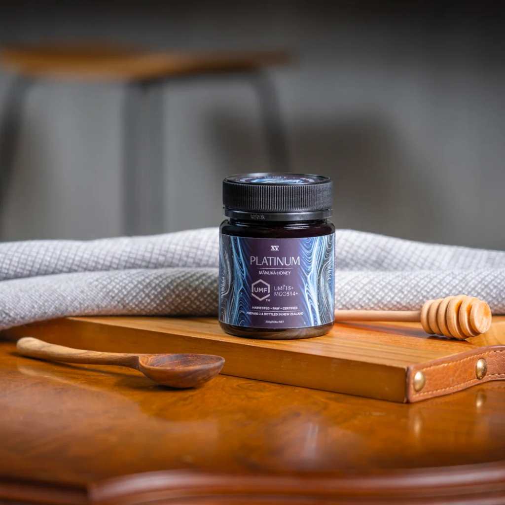 Jar of platinum Manuka Honey on a wooden surface with a honey dipper and cloth in the background