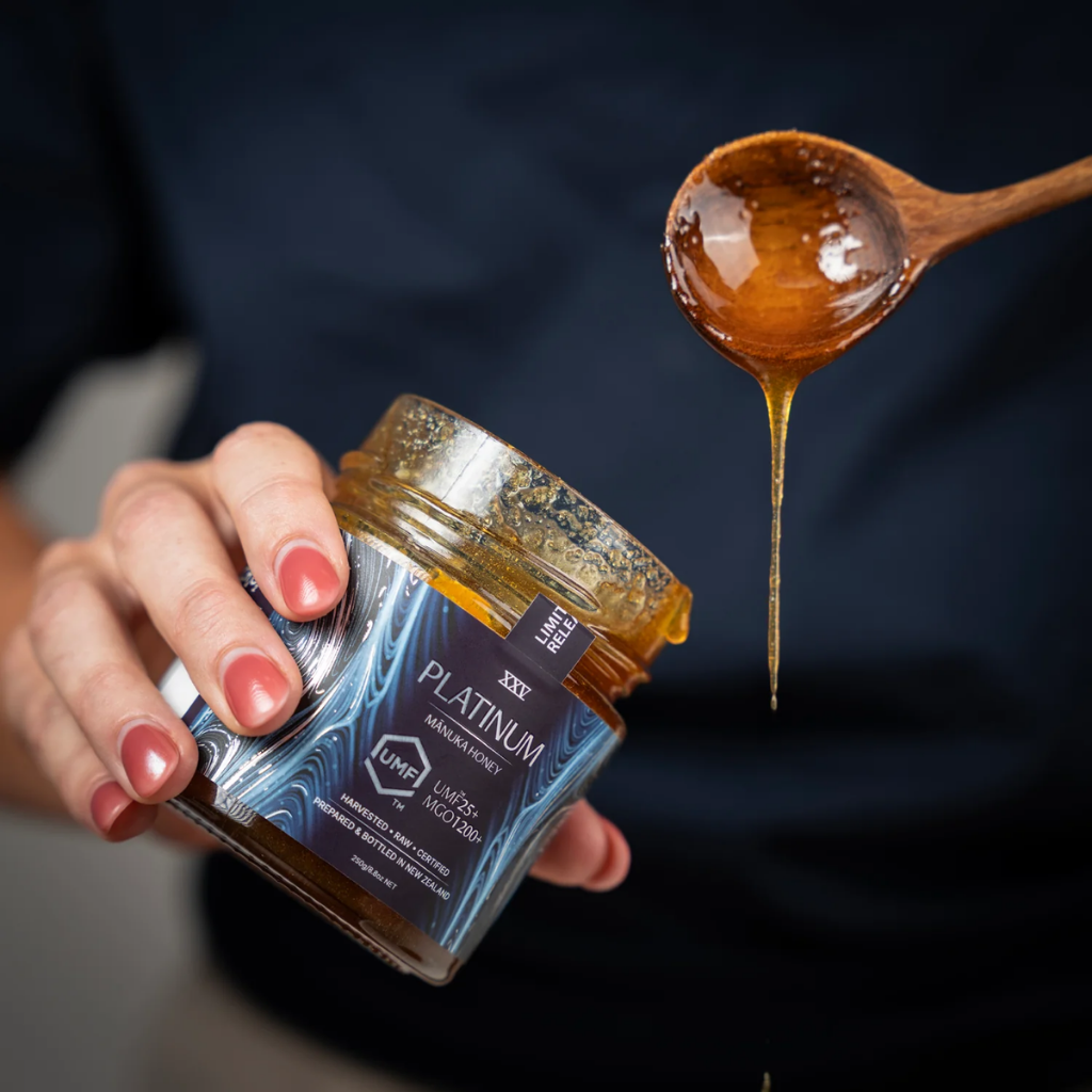 Hand holding a jar of honey labeled 'Platinum' with a wooden spoon dripping honey above.