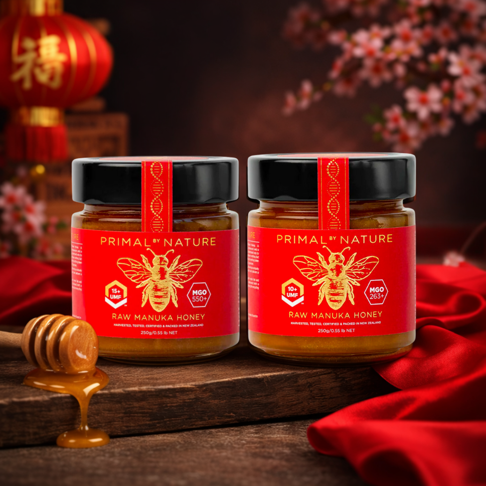 Two jars of Primal Nature Raw Manuka Honey on a wooden surface with a red cloth and honey dipper in the background.