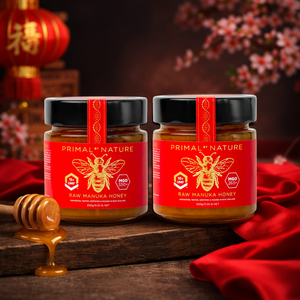 Two jars of Primal Nature Raw Manuka Honey on a wooden surface with a red cloth and honey dipper in the background.