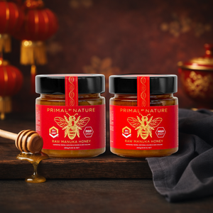 Two jars of Primal Nature raw manuka honey on a wooden surface with a honey dipper.