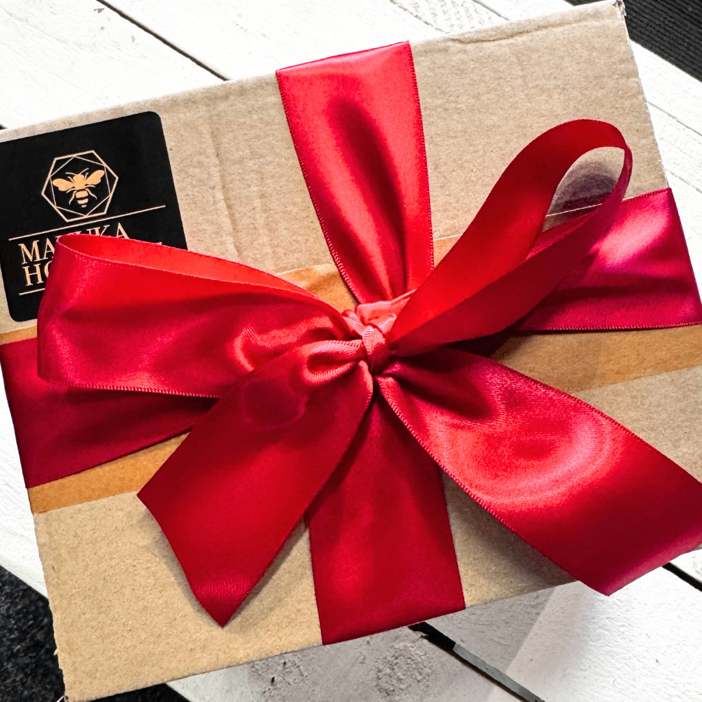 A square kraft paper gift box wrapped with a large shiny red satin ribbon bow and tied with a matching red ribbon, featuring a black brand label on the top left.