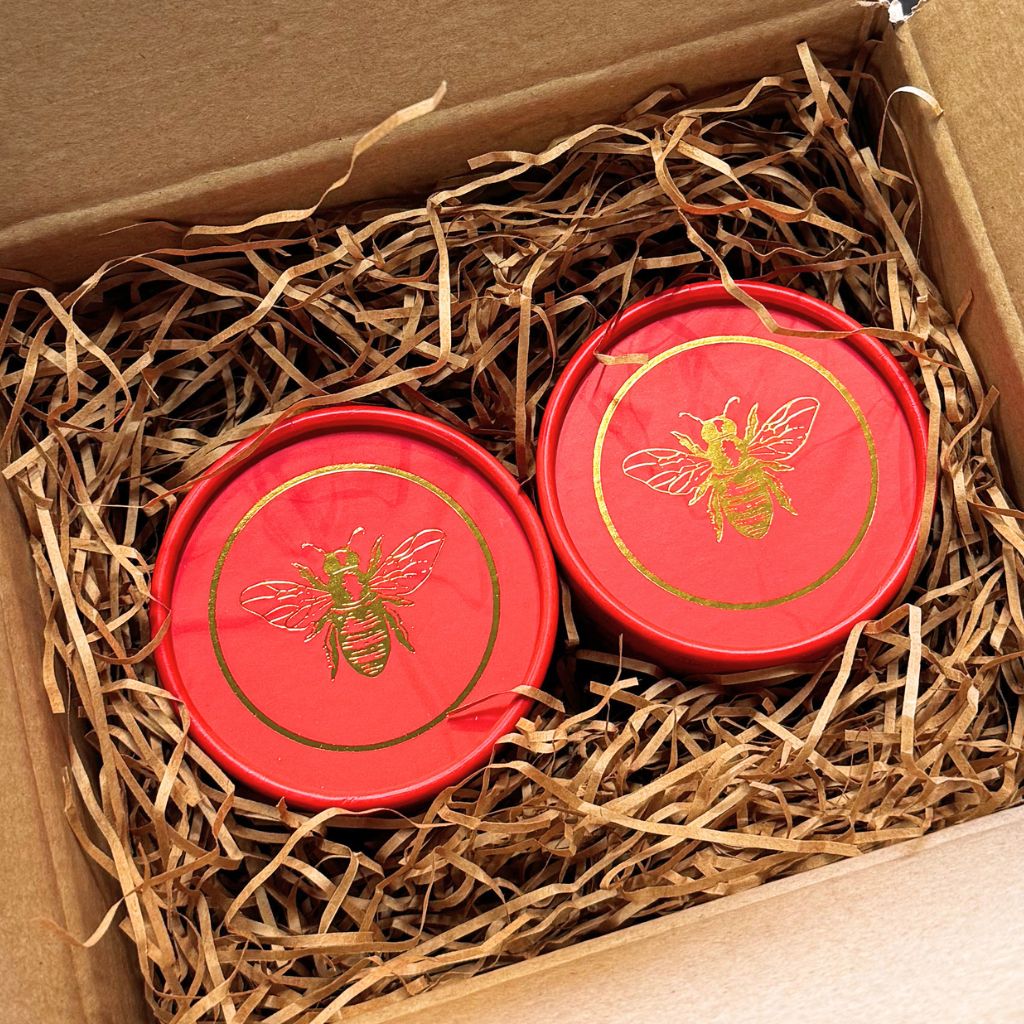 Two round red gift boxes with gold bee designs nestled in brown paper shred inside a cardboard box.