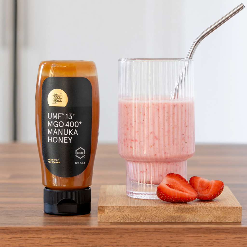 Manuka honey bottle next to a glass of strawberry smoothie on a wooden surface