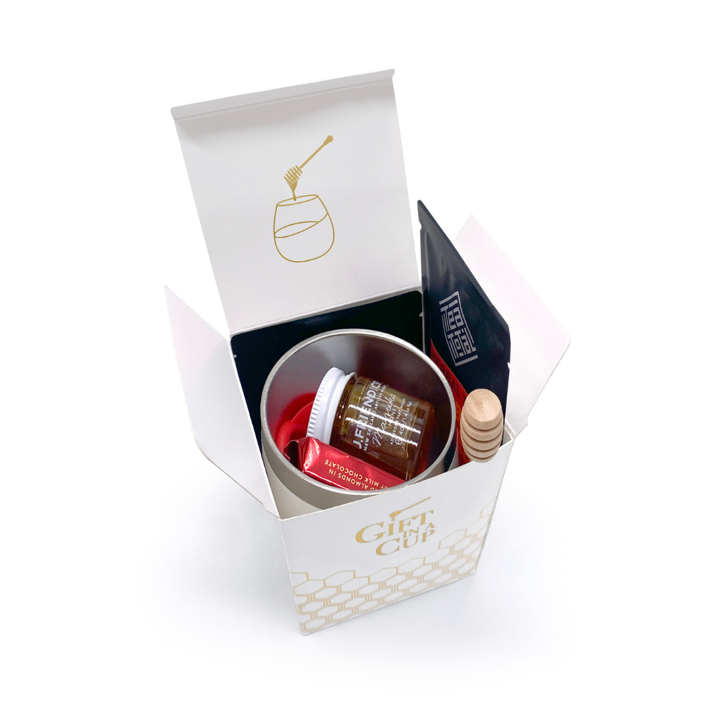 A collection of gift items including a white tumbler with a lid, a jar of manuka honey, loose leaf tea packets, a red silicone tea strainer, a wooden honey dipper, and a small bar of chocolate, all arranged with Christmas decorations in the background.