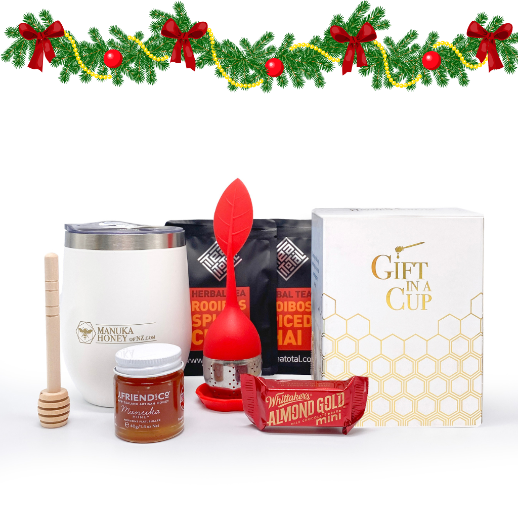A collection of gift items including a white tumbler with a lid, a jar of manuka honey, loose leaf tea packets, a red silicone tea strainer, a wooden honey dipper, and a small bar of chocolate, all arranged with Christmas decorations in the background.