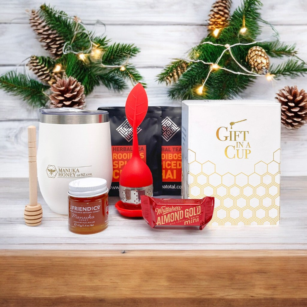 A collection of gift items including a white tumbler with a lid, a jar of manuka honey, loose leaf tea packets, a red silicone tea strainer, a wooden honey dipper, and a small bar of chocolate, all arranged with Christmas decorations in the background.