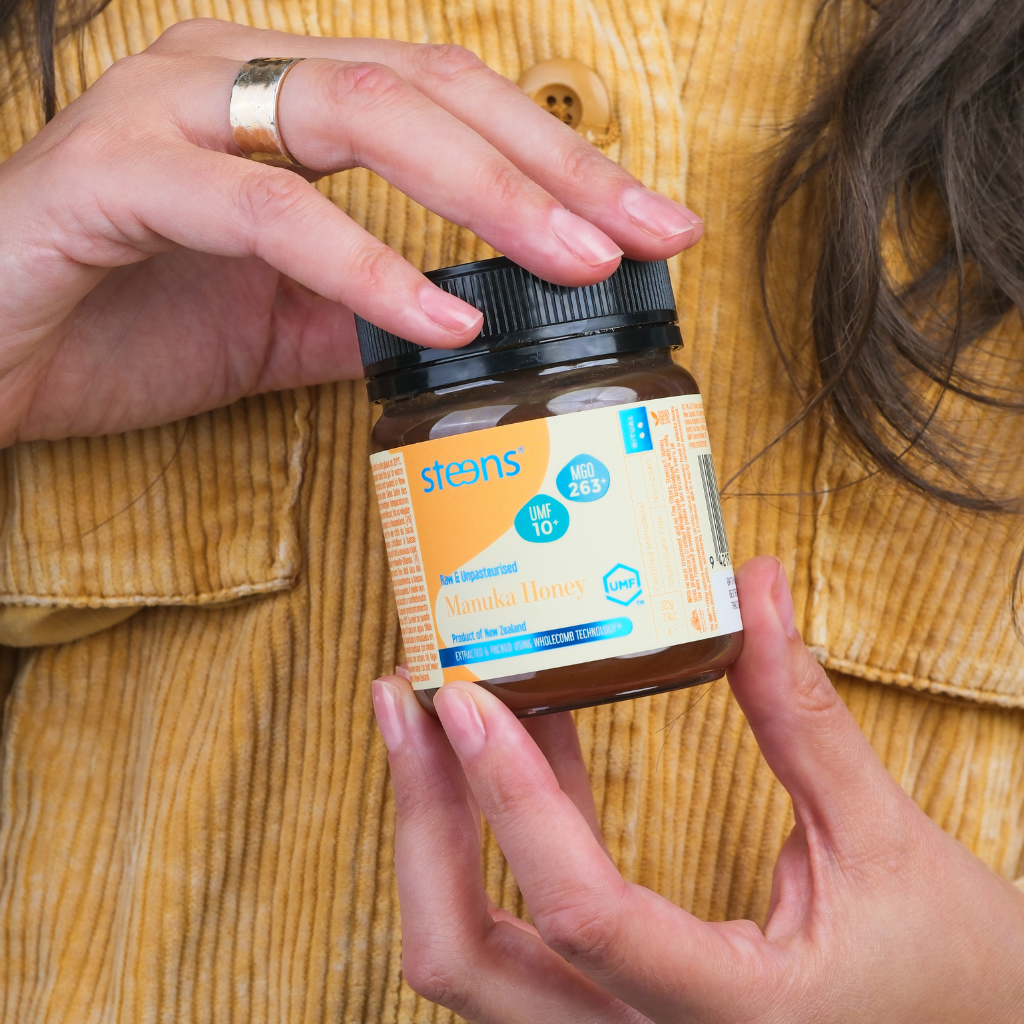 A person holding a jar of Steens Manuka Honey against a yellow corduroy shirt background. The jar has a black lid and a cream-colored label with blue and orange text.