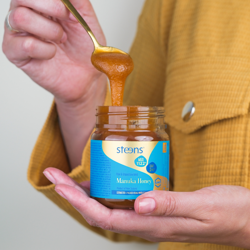 A person in a mustard yellow jacket holds an open jar of Steens Mamika Honey while lifting honey with a gold spoon.