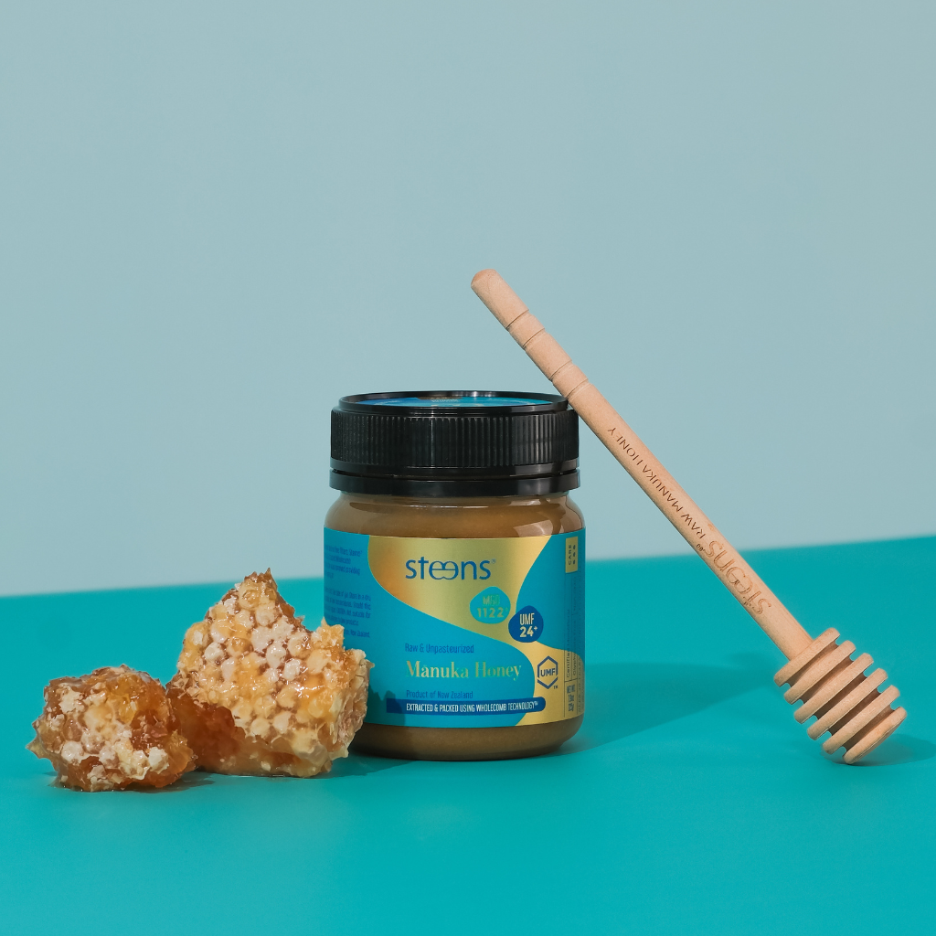 A jar of Steens Manuka Honey with a black lid and blue label, styled with two pieces of raw honeycomb and a wooden honey dipper on a turquoise background.