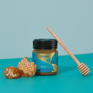 A jar of Steens Manuka Honey with a black lid and blue label, styled with two pieces of raw honeycomb and a wooden honey dipper on a turquoise background.